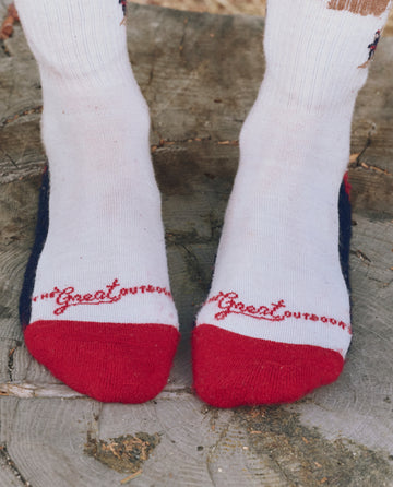 A pair of feet in THE GREAT. F25 TGO The Ridge Runner Sock -- Cream Skiier, cream merino wool socks with red toes and "Great Outdoors" near the toes, standing on a tree stump.