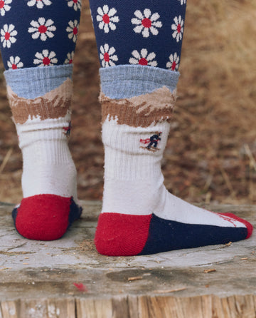A person stands on a tree stump in daisy leggings and THE GREAT. F25 TGO The Ridge Runner Sock—Cream Skiier, featuring a mountain landscape in red, blue, and white with a cushioned footbed. Pine needles cover the ground below.