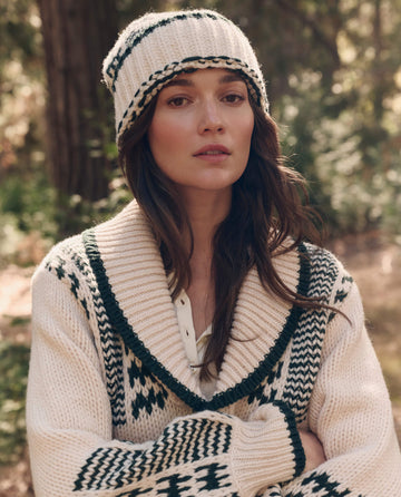 A woman stands in a wooded area wearing THE GREAT. F26 TGO The Chalet Beanie. -- Cream And Forest, looking at the camera with a neutral expression.