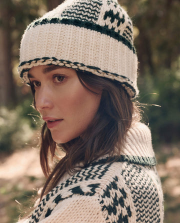 A woman wearing The Chalet Beanie. -- Cream And Forest by THE GREAT. F26 TGO stands outdoors in a matching sweater, looking to the side with a neutral expression.