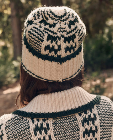 Someone stands outdoors facing away, wearing THE GREAT. F26 TGO The Chalet Beanie. -- Cream And Forest with a matching sweater.