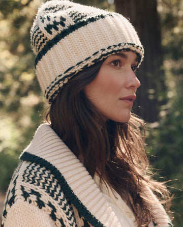 A woman wearing THE GREAT. F26 TGO The Chalet Beanie. -- Cream And Forest stands outdoors in a wooded area, looking to the side.