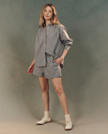 A person stands in a relaxed pose wearing THE GREAT. SP25 D2's The Boxer Short in Black and White Gingham, paired with white socks and shoes, against a neutral background.