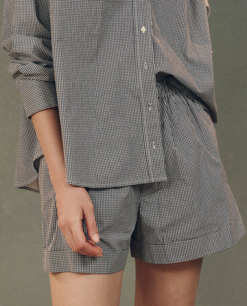A person models "The Boxer Short. -- Black And White Gingham" by THE GREAT. SP25 D2, with the long-sleeve shirt tucked slightly into matching boxer shorts, set against a plain background, illustrating a comfortable fit.