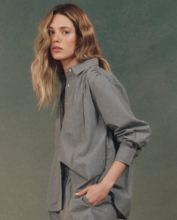 Against a neutral backdrop, a long-haired person with wavy locks poses with one hand in their pocket while wearing THE GREAT. SP25 D2 "The Society Top" in black and white gingham—a stylish checkered shirt and pants.