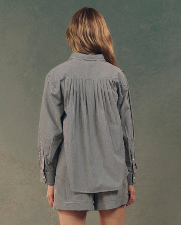 A person with long hair, seen from behind, wears The Society Top in black and white gingham with matching shorts against a muted green background. Brand: THE GREAT. SP25 D2.