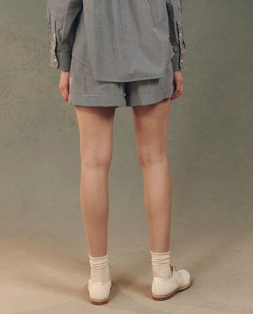 Rear view of a person in a striped long-sleeve shirt, THE GREAT. SP25 D2 The Boxer Short in Black and White Gingham, white socks, and cream shoes against a neutral background for a comfortable fit.