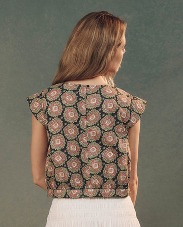 A woman with long hair faces away, wearing The Midland Top in Heirloom Paisley by THE GREAT. SP25 D2, paired with a high-waisted white skirt against a plain backdrop.