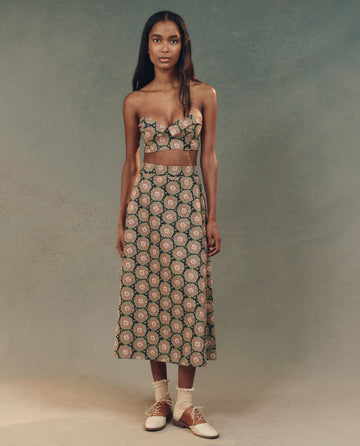 In a studio, an elegant fashion display features THE GREAT. SP25 D2's Heirloom Paisley Bustier Top with a floral two-piece outfit and neutral shoes, complementing the soft green and beige gradient background.