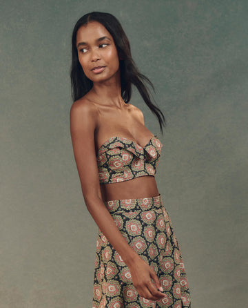 A woman in an elegant ensemble featuring THE GREAT. SP25 D2's The Bustier Top in Heirloom Paisley and a matching skirt, looks to the side against a textured background.