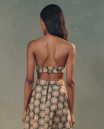 A person with long dark hair stands gracefully with their back to the camera, wearing an elegant ensemble featuring THE GREAT. SP25 D2 Heirloom Paisley Bustier Top and matching pants.