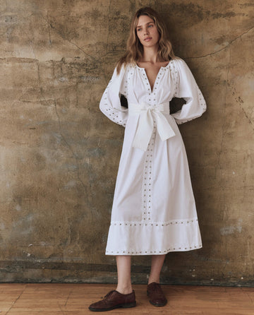 A woman in THE GREAT. SP25 POPLIN's The Bodega Dress with Studs in white stands by a textured brown wall, arms relaxed. She completes the elegant look with brown studded shoes on a wooden floor.