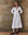 A woman in THE GREAT. SP25 POPLIN's The Bodega Dress with Studs in white stands by a textured brown wall, arms relaxed. She completes the elegant look with brown studded shoes on a wooden floor.