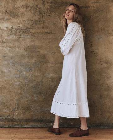 A person in a long white "The Bodega Dress with Studs" by THE GREAT. SP25 POPLIN and brown shoes leans against a textured brown wall, glancing over their shoulder.