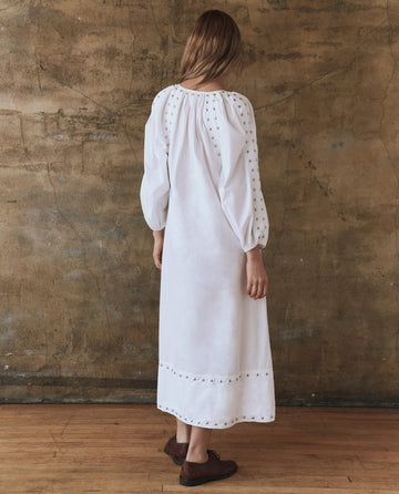 A person wearing The Bodega Dress with Studs in white by THE GREAT. SP25 POPLIN stands facing a textured, brownish wall on a wooden floor.