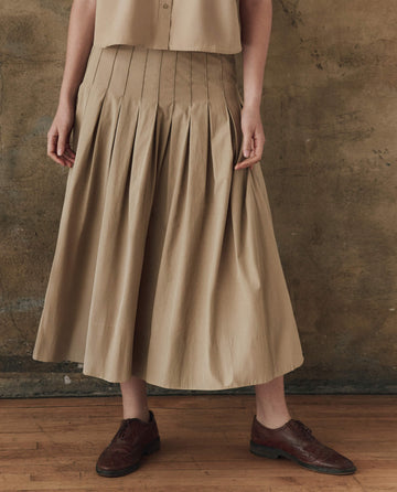 Wearing THE GREAT. SP25 POPLIN matching set, a person stylishly models The Rhythm Skirt in khaki, standing with hands at their sides on a wooden floor against a textured wall.