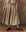 Wearing THE GREAT. SP25 POPLIN matching set, a person stylishly models The Rhythm Skirt in khaki, standing with hands at their sides on a wooden floor against a textured wall.