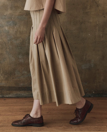 Wearing The Rhythm Skirt in khaki from THE GREAT. SP25 POPLIN, a person walks gracefully on a wooden floor against a textured brown wall, their steps harmonizing with the skirt's timeless elegance.