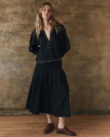 A person wearing The Rhythm Skirt in black from THE GREAT. SP25 POPLIN stands against a textured brown wall. Their brown shoes enhance the understated elegance of this fashionable ensemble, with arms gracefully placed behind their back.