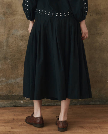 A person in THE GREAT. SP25 POPLIN's stylish black Rhythm Skirt pairs it with a patterned top, standing elegantly on a wooden floor against a textured wall.