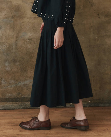 A person stands against a textured wall wearing a black, long-sleeved shirt with beaded embellishments, The Rhythm Skirt in Black by THE GREAT. SP25 POPLIN, and brown leather brogues.