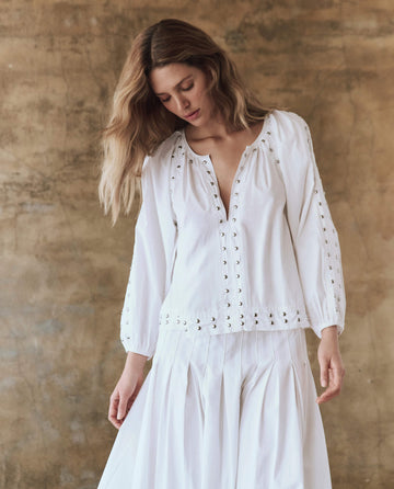 A person with long hair models The Bodega Top with Studs from THE GREAT. SP25 POPLIN, paired with a chic skirt against a beige wall, exuding modern fashion vibes and enhanced by the top's subtle studded allure as they gaze downward.