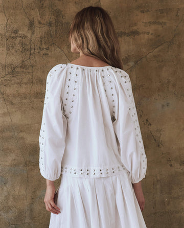 Wearing THE GREAT.'s white Bodega Top with Studs, a chic individual with long hair poses gracefully against a textured brown wall, exuding effortless style. The subtle studs add an edgy touch to the ensemble's understated charm.