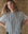 Wearing The Valor Top in Parchment Stripe by THE GREAT. SP25 POPLIN, a person stands confidently with arms akimbo against a textured backdrop, highlighting the sophisticated style of women's fashion.