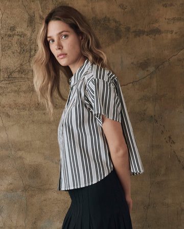 A person with long hair stands against a textured wall, wearing The Valor Top in Parchment Stripe by THE GREAT. SP25 POPLIN and dark pants, looking at the camera.