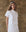 A woman in a white button-up from THE GREAT. SP25 POPLIN's The Valor Top collection and a matching skirt stands against a textured brown wall with a striped headband.