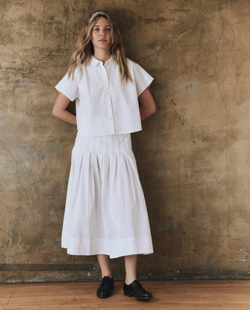 A person with long hair, dressed in a stylish white The Valor Top by THE GREAT. SP25 POPLIN and a skirt, stands against a textured brown wall wearing black shoes.