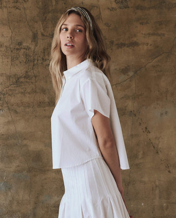 A person with long hair stands against a textured brown wall, wearing The Valor Top in white by THE GREAT. SP25 POPLIN and a white skirt, showcasing the elegance of women's clothing.