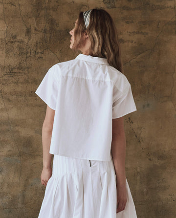 A person with long hair, wearing THE GREAT. SP25 POPLIN's chic white Valor Top and a matching skirt, stands fashionably facing away in front of a textured brown wall.