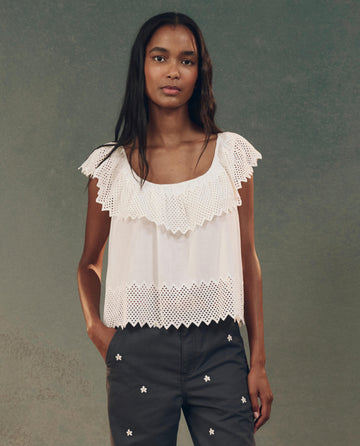 A woman in a white, sleeveless Westport Top by THE GREAT. SP25 D2, featuring honeycomb eyelet details, stands against a neutral background wearing dark pants adorned with small floral patterns.