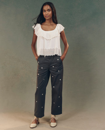 A woman stands against a neutral background wearing the white "The Westport Top with Honeycomb Eyelet" by THE GREAT. SP25 D2, paired with dark pants featuring small white designs and stylish light-colored shoes.