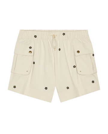 The Embroidered Cargo Short by THE GREAT. in cream features black daisy embroidery, an elastic waistband with front drawstring, and two large flap pockets.