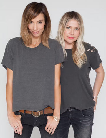 Two women with light brown and blonde hair wearing casual grey t-shirts and jeans stand together against a plain background.