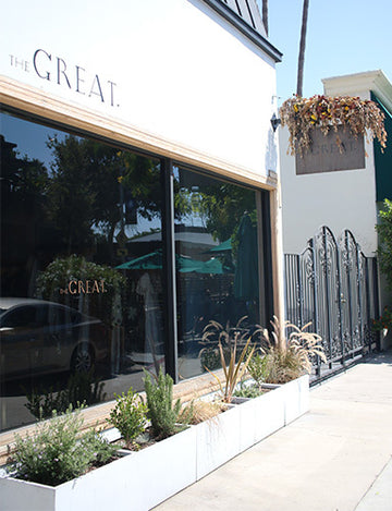 Street view of a store with a sign reading "The Great." The storefront features large glass windows and potted plants in front. A hanging sign and decorative gate are on the right.
