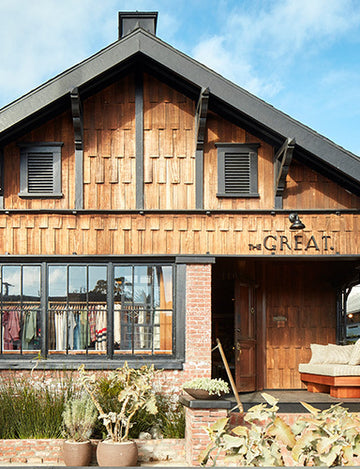 A rustic two-story wooden building with a brick base and large windows. A sign reads "THE GREAT." Outdoor seating and potted plants are visible.