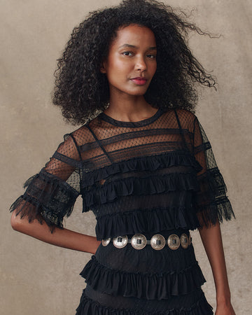A woman with curly hair wearing a black sheer ruffled dress with a silver belt stands in front of a plain, beige background.