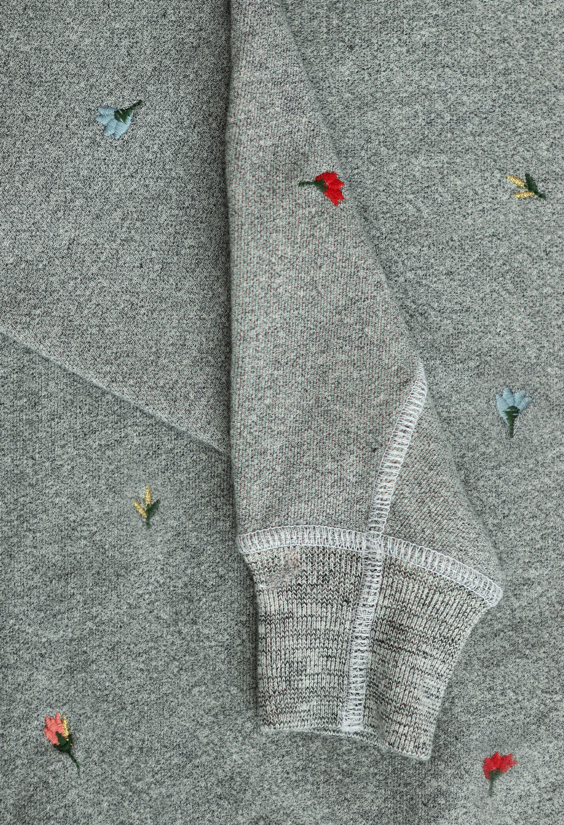 Close-up of a gray sweatshirt with small embroidered flowers in red, blue, and yellow, and a visible folded sleeve.