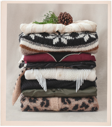 A neatly folded stack of sweaters in various colors and patterns, topped with greenery and a pine cone.