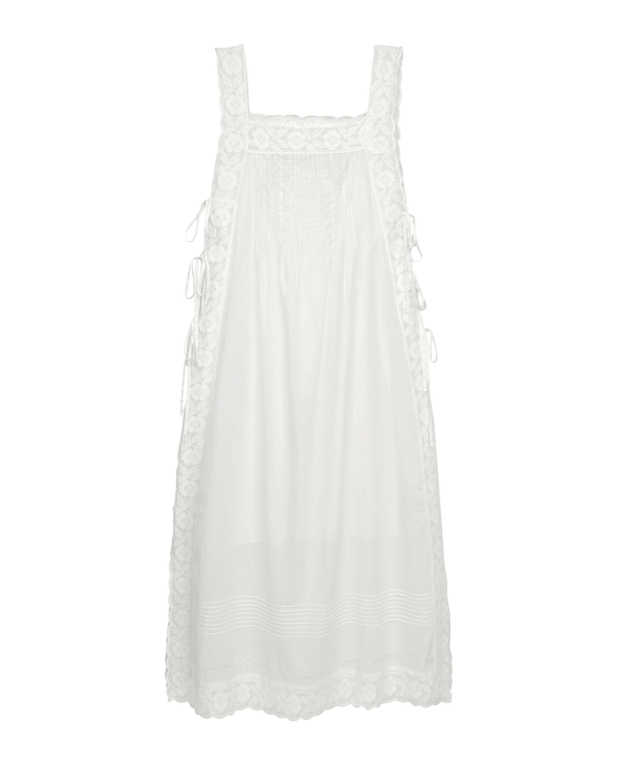 The Attic Lace Dress by THE GREAT. SU25 in white features lace trim, a square neckline, side ties, and front vertical pleats, displayed on a black background.