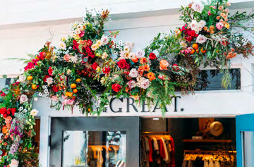 A storefront adorned with a vibrant floral arrangement above the entrance, featuring red, pink, and orange flowers, alongside green foliage. Visible clothing inside through the glass doors.