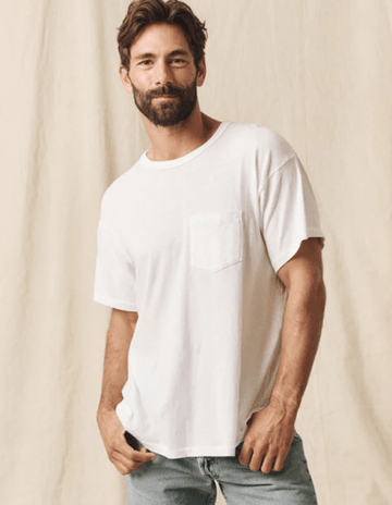 A man with a beard wearing a white t-shirt and jeans stands against a beige background.