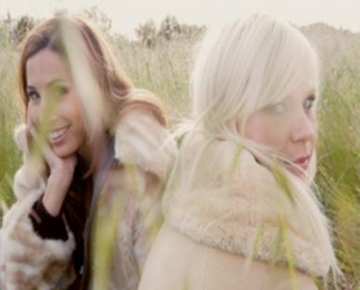 Two people in fur coats sit in a grassy field. One person has dark hair and is smiling, while the other has light hair and a neutral expression.