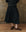 A person in a black THE GREAT. SP25 POPLIN Rhythm Skirt, a studded black top, and brown brogues stands on a wooden floor against a textured brown wall, effortlessly highlighting their unique fashion sense.