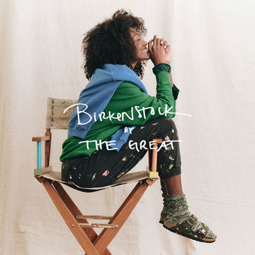 Person with curly hair sits sideways on a wooden chair, wearing layered clothing and sandals. Text overlay reads "Birkenstock by The Great.