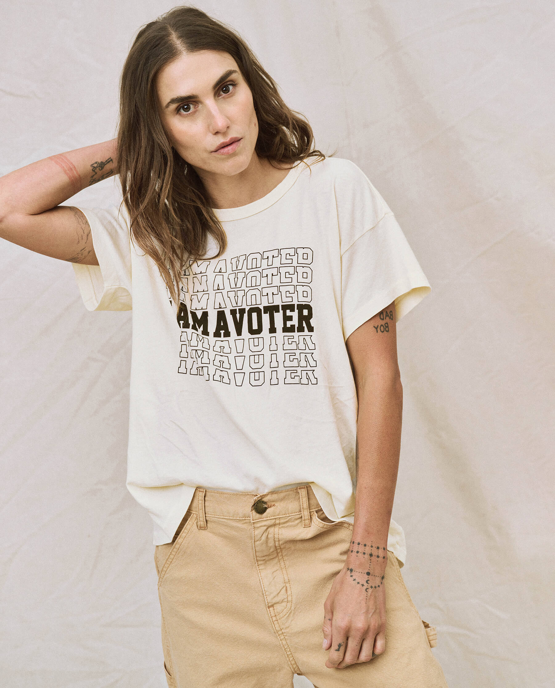 A person with long hair stands against a light fabric backdrop, wearing the limited edition "The I Am A Voter Boxy Crew" from The Great. in washed white, featuring repeated "I AM A VOTER" text, paired with beige pants. Their left arm is raised to their head, highlighting the casual style of this unique tee.