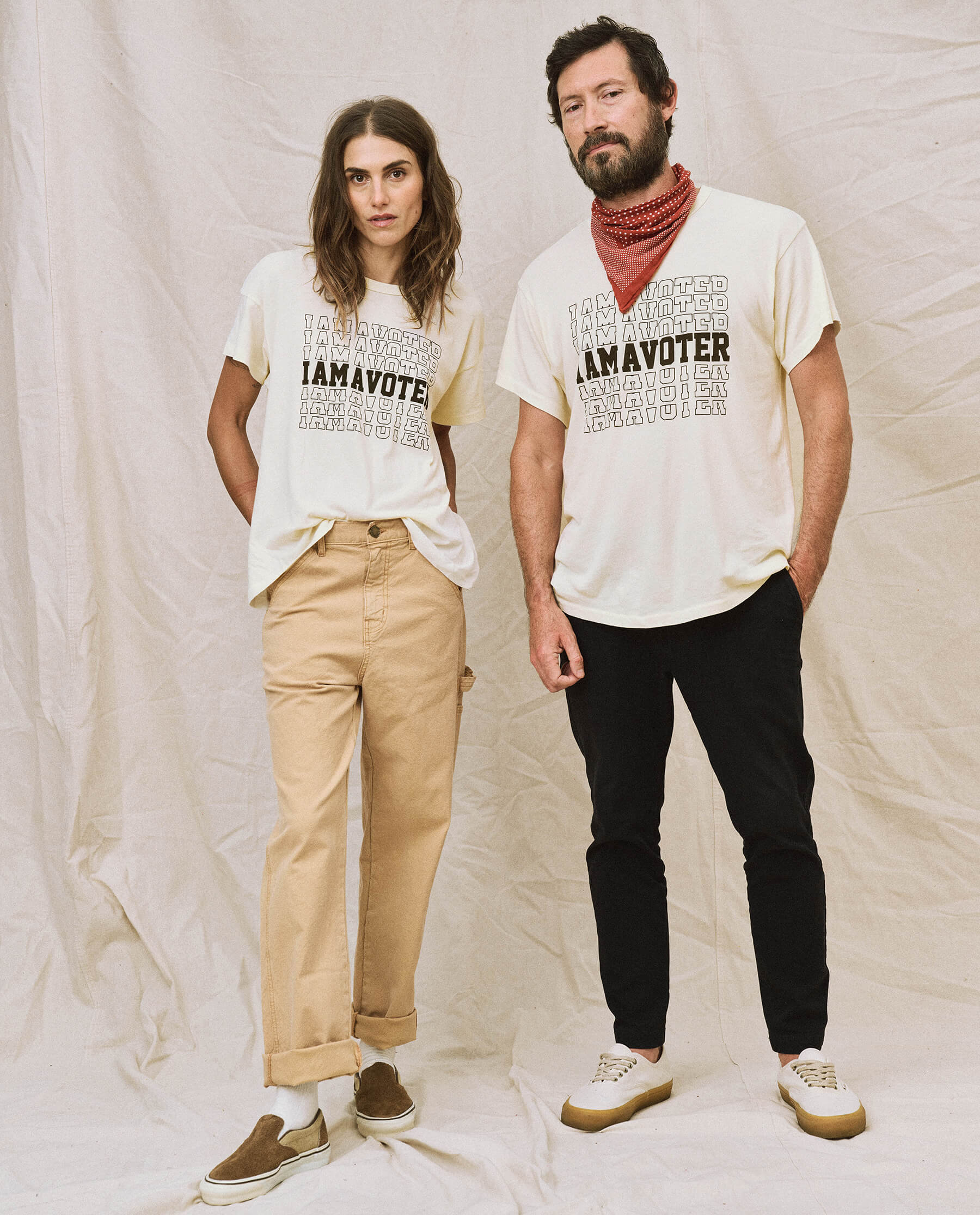 Two people wearing the limited edition "The I Am A Voter Boxy Crew" T-shirts by The Great. in washed white stand in front of a beige backdrop; the person on the left wears beige pants, while the person on the right pairs black pants with a red bandana.